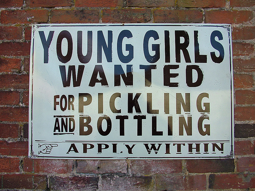 A white sign with black text is mounted to a red brick wall. The sign reads: "Young girls wanted for pickling and bottling. Apply within." The sign's bad grammar, of course, leaves open the interpretation to believe they want to pickle young girls and then bottle them.